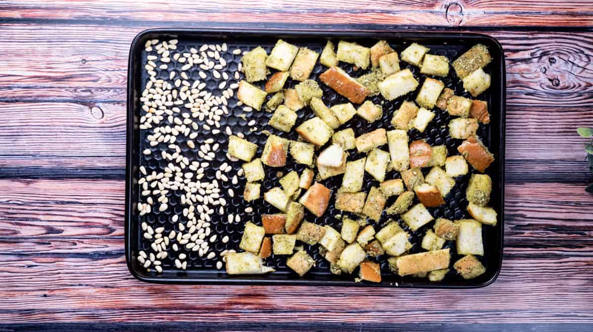 A baking tray with pesto bread cubes and pine nuts laid out on it.