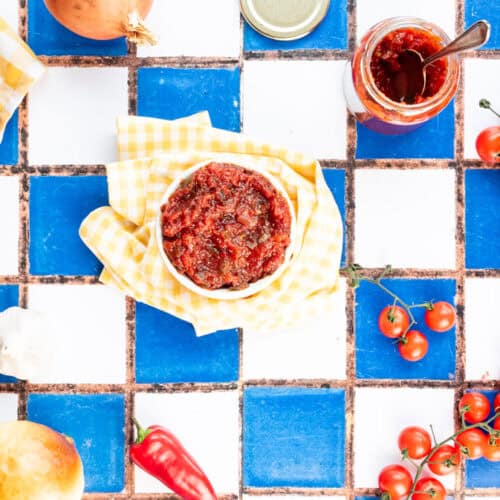 A bowl of tomato relish surrounded by ingredients, decorative items, and jars of relish.