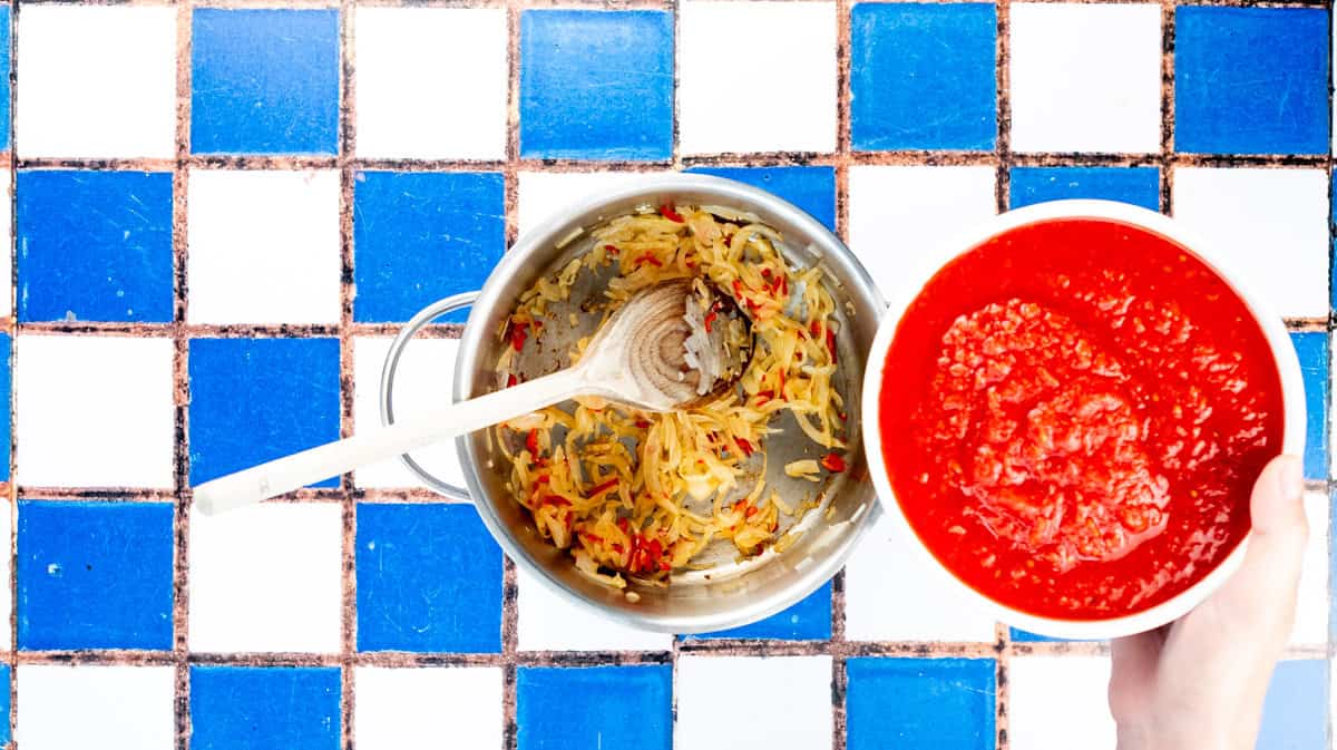 A hand is pouring chopped tomatoes in saucepan with the cooked vegetables inside.
