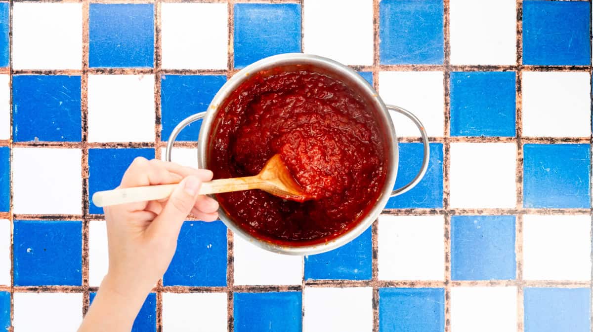 A hand is using a wooden spoon to stir the burger relish in a saucepan as it thickens.