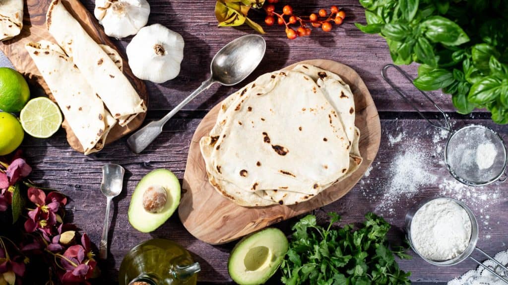 An overhead view of a pile of tortillas wraps on a wooden board. There are some rolled up next next to them and the wraps are surrounded by ingredients, like avocado, olive oil, limes, fresh herbs and garlic.