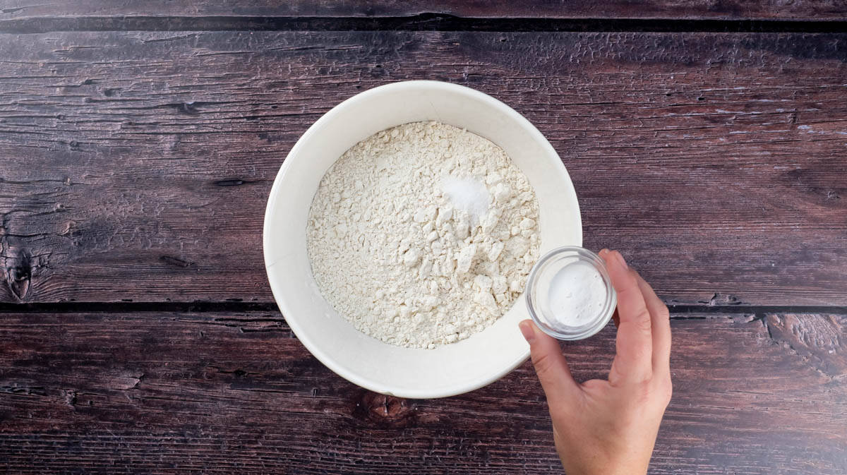 A hand is adding baking powder to a bowl of flour with added salt.
