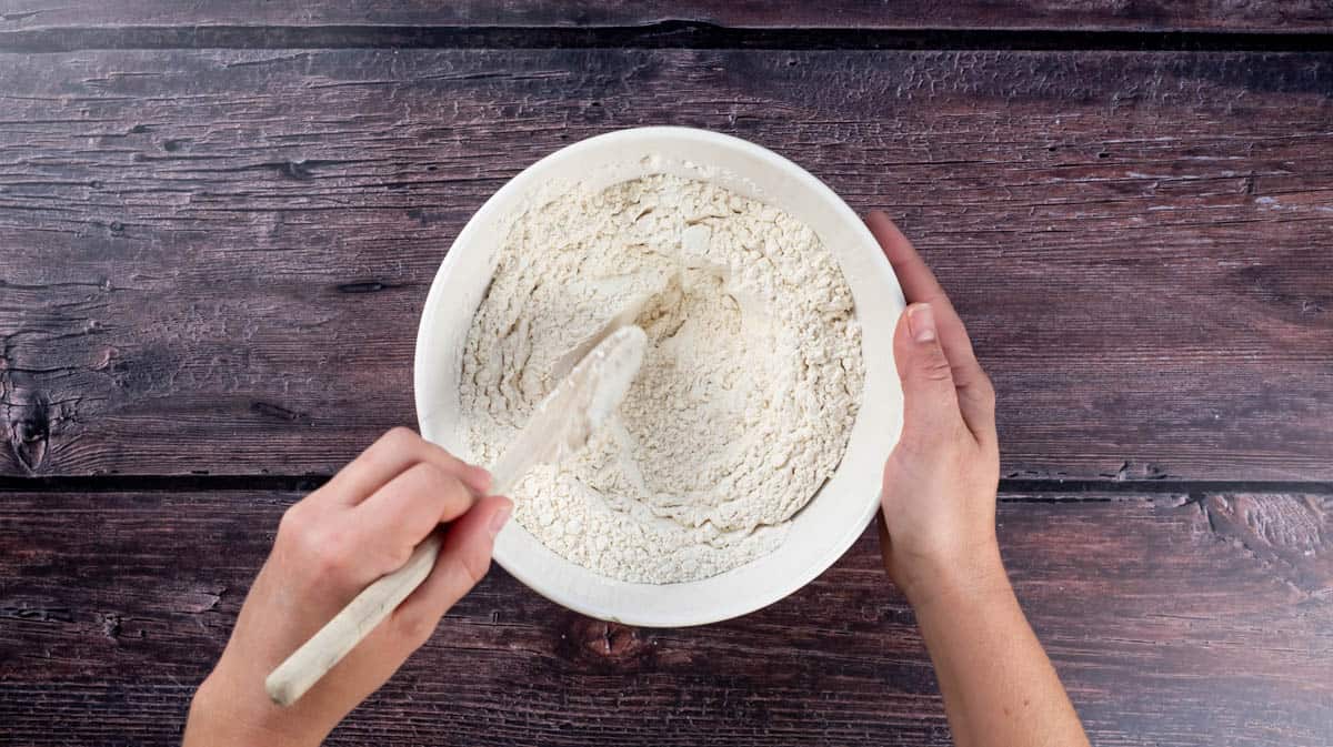 A hand is using a wooden spoon to mix the flour mixture.
