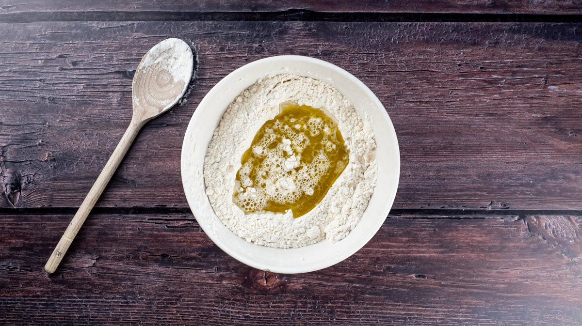 An overhead view of a bowl of flour mixture with added water and olive oil in the centre.