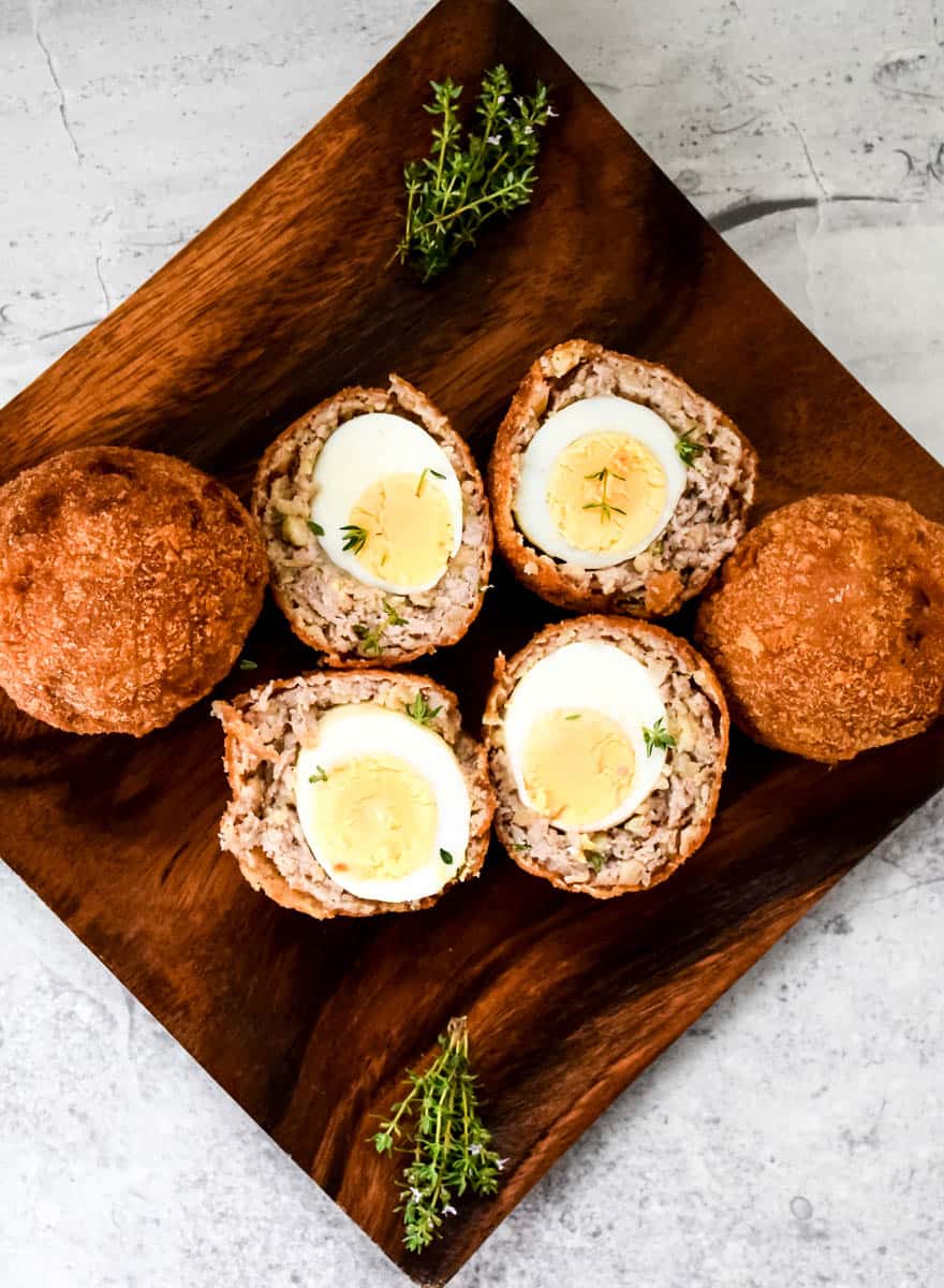 A square wooden plate with four Scotch eggs and a bunch of thyme. 2 Scotch eggs have been sliced in half and the other two remain whole.