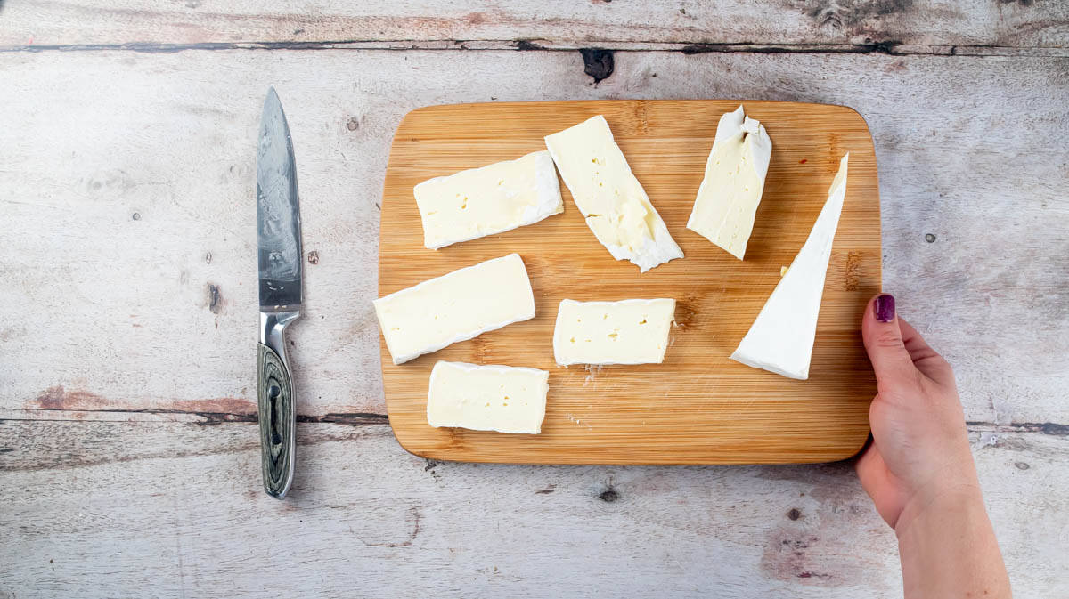 A hand is holding a wooden board containing slices of brie cheese next to a knife.