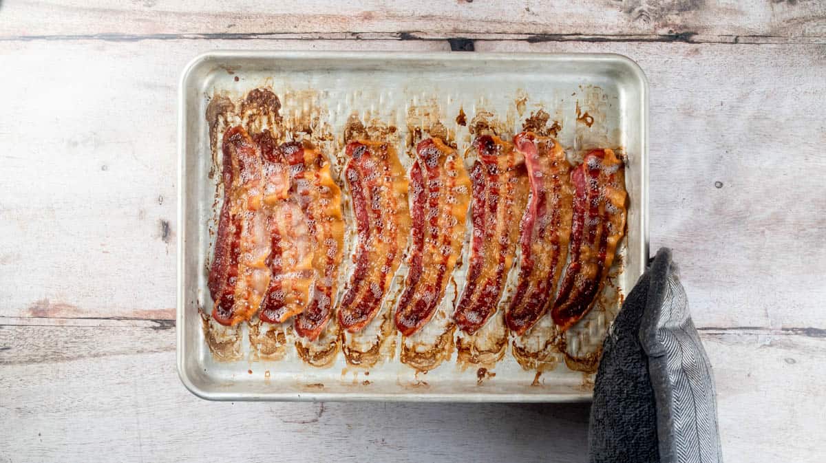 A gloved hand is holding a silver baking tray with eight cooked slices of bacon upon it.
