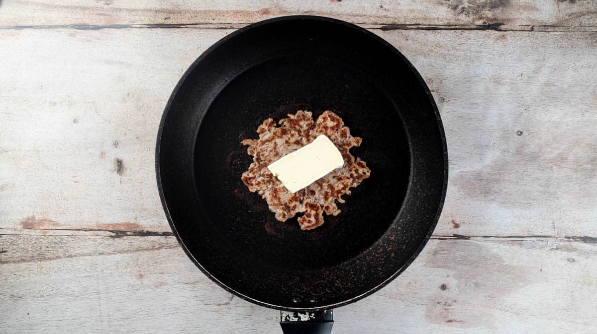 A finished burger patty in the frying pan with a slice of brie cheese on top.