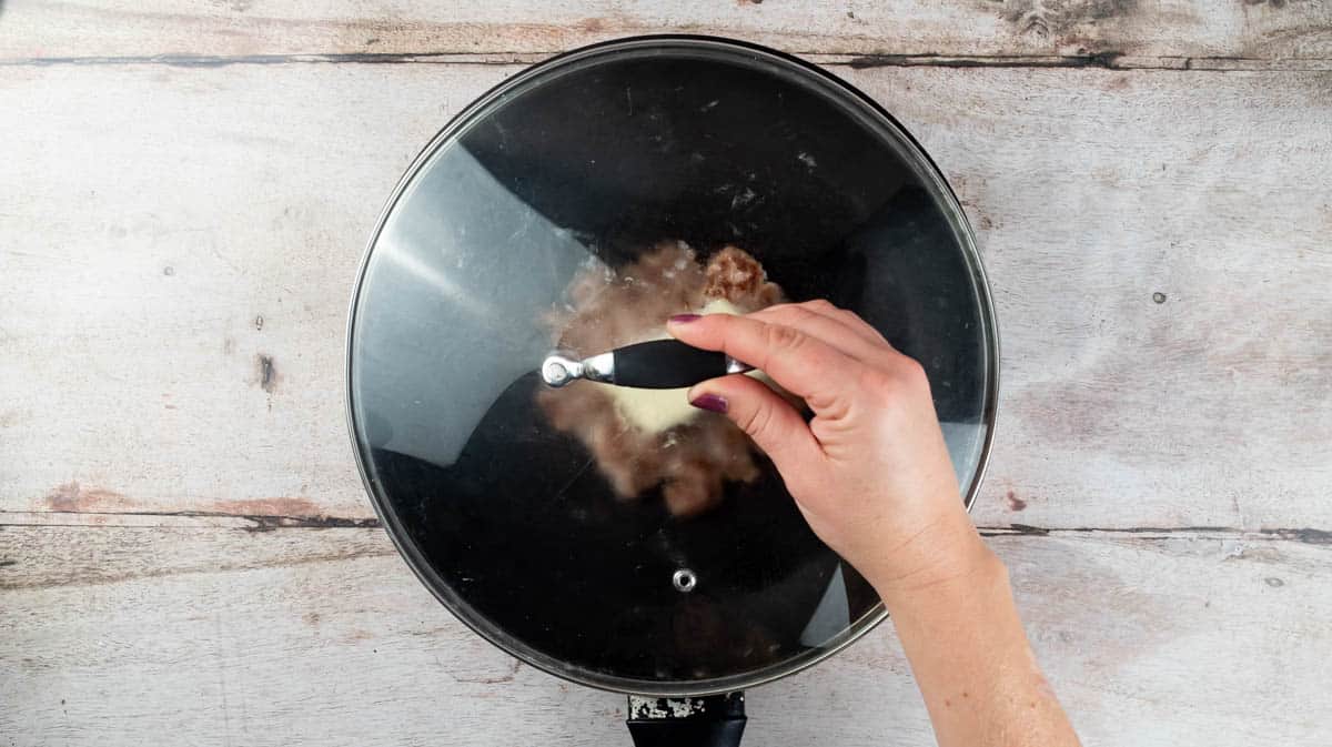 A hand is closing the top of the frying pan with a lid to melt the cheese.