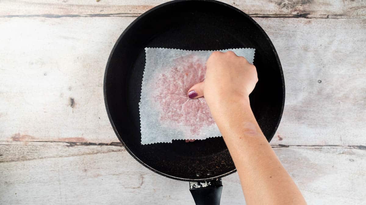 A hand is pressing a thumb into the middle of the smash burger that is still covered with baking paper.