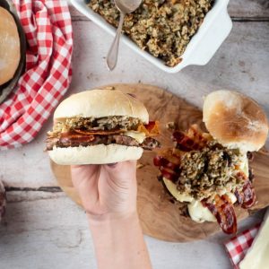 A hand is holding the completed turkey smash burger up to the camera to show the filling. Behind is another burger on a wooden board, red decorative items and ingredients.