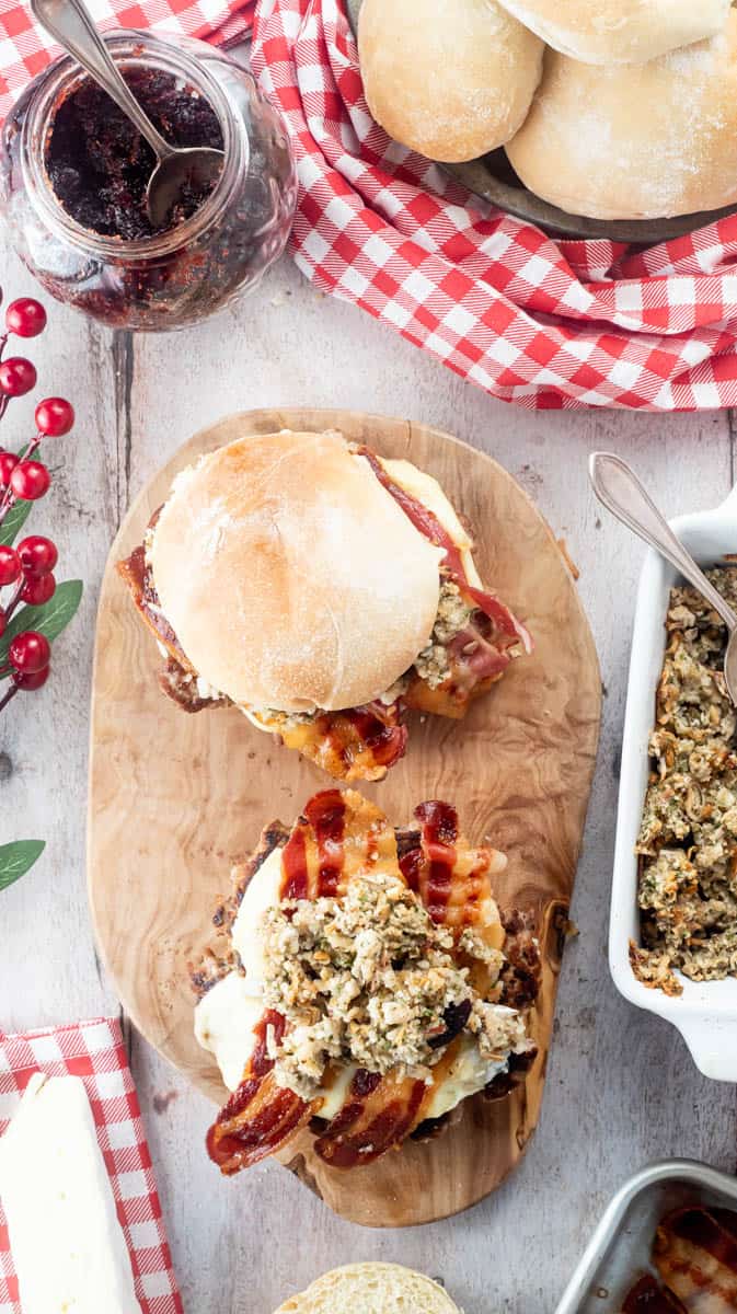 An overhead view of two turkey burgers, one is closed and the other is open to show the filling. They are sat on a wooden board surrounded by red themed decorations and ingredients.