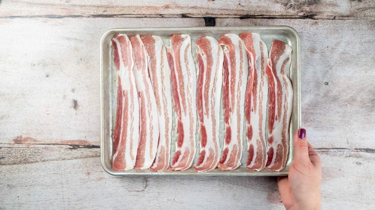 A hand is holding a silver baking tray containing eight slices of streaky bacon uncooked.