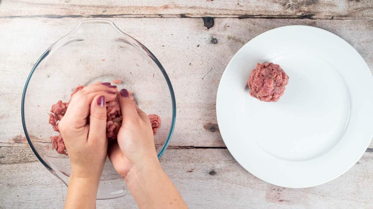 Two hands are shaping some turkey mince into a ball next to a white plate that already contains one completed ball.