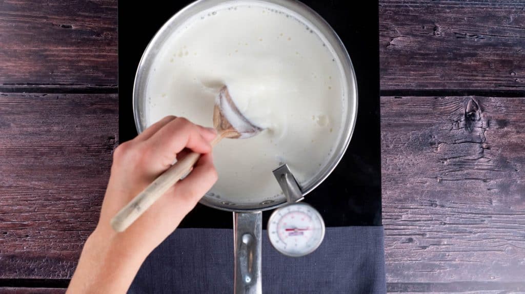 A hand is stirring the milk as it heats with a thermometer inside it.